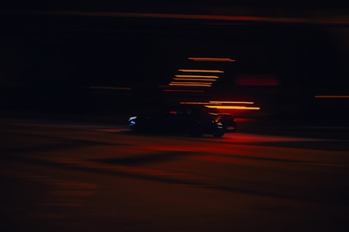 A car moving at high speed is captured with motion blur, creating a dynamic sense of motion. The image is dimly lit, with streaks of red and orange lights appearing against a dark background.