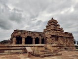 An ancient stone temple with intricate carvings and a towering structure against a backdrop of overcast skies. The temple is built with sandstone and features detailed architectural elements, such as columns and pillars. The surrounding area is clear, emphasizing the temple's prominence and historical significance.