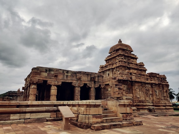 An ancient stone temple with intricate carvings and a towering structure against a backdrop of overcast skies. The temple is built with sandstone and features detailed architectural elements, such as columns and pillars. The surrounding area is clear, emphasizing the temple's prominence and historical significance.