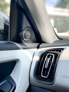 A close-up view of a car interior featuring a speaker and an air vent on the dashboard. The speaker is located near the side mirror and has a circular design with a grille pattern. The air vent has a rectangular shape with a metallic frame. The materials appear to be a mix of leather and plastic, contributing to a modern and clean aesthetic.