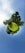 A surreal miniature planet landscape with lush green grass and dense trees forming a circular shape against a vast blue sky. The scene appears as if a small, grassy globe is floating in the sky, with people visible in the center enjoying the outdoors.