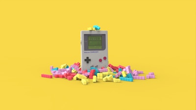 A vintage handheld gaming console surrounded by colorful, block-like pieces reminiscent of a classic puzzle game. The console has a small screen displaying a game in progress, with two red buttons and a directional pad. The vibrant pieces are scattered around it, creating a playful and nostalgic atmosphere.