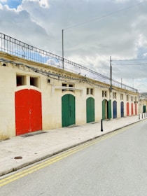 A row of colorful garage doors is set against a pale stone building. The doors are painted in various colors, including red, green, and blue. Overhead, a wrought iron railing lines the roof. The sky is mostly cloudy, casting a soft light on the scene. The street is empty, with double yellow lines and a row of black bollards lining the sidewalk.