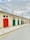 A row of colorful garage doors is set against a pale stone building. The doors are painted in various colors, including red, green, and blue. Overhead, a wrought iron railing lines the roof. The sky is mostly cloudy, casting a soft light on the scene. The street is empty, with double yellow lines and a row of black bollards lining the sidewalk.