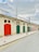 A row of colorful garage doors is set against a pale stone building. The doors are painted in various colors, including red, green, and blue. Overhead, a wrought iron railing lines the roof. The sky is mostly cloudy, casting a soft light on the scene. The street is empty, with double yellow lines and a row of black bollards lining the sidewalk.