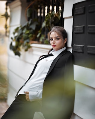 A person in a suit leans against an outdoor wall, surrounded by greenery. The expression suggests confidence and calmness. The background includes blurred foliage, adding depth to the image.