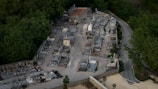 An aerial view of a cemetery surrounded by dense green trees. The cemetery contains several rows of graves with headstones and small mausoleums. A narrow road runs alongside the cemetery, and a small chapel or building with a red roof is positioned near the center.