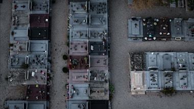 An aerial view of a cemetery features rows of graves with various tombstones, ranging from simple to more elaborate designs. Many plots have flowers, vases, and decorative items placed on them. The ground between the graves is covered in gravel and there are small patches of greenery.