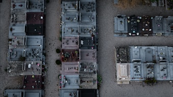 An aerial view of a cemetery features rows of graves with various tombstones, ranging from simple to more elaborate designs. Many plots have flowers, vases, and decorative items placed on them. The ground between the graves is covered in gravel and there are small patches of greenery.