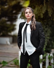 A person stands confidently in a black suit with a white shirt and black tie, set against a blurred outdoor background with greenery.