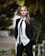 A person stands confidently in a black suit with a white shirt and black tie, set against a blurred outdoor background with greenery.