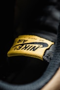A close-up view of a yellow fabric label with the word 'Nike' and the swoosh logo, stitched onto a black surface. The texture of the fabric and stitching details are clearly visible.