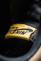 A close-up view of a yellow fabric label with the word 'Nike' and the swoosh logo, stitched onto a black surface. The texture of the fabric and stitching details are clearly visible.