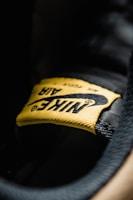 A close-up view of a yellow fabric label with the word 'Nike' and the swoosh logo, stitched onto a black surface. The texture of the fabric and stitching details are clearly visible.