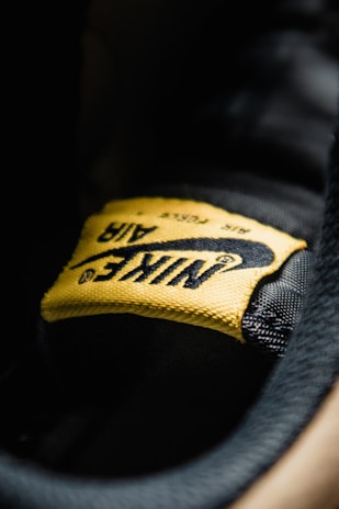 A close-up view of a yellow fabric label with the word 'Nike' and the swoosh logo, stitched onto a black surface. The texture of the fabric and stitching details are clearly visible.