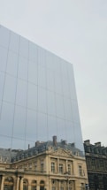 Reflection of a historic building with ornate architecture on the mirrored surface of a modern glass structure. The reflected building features classical elements such as columns and decorative moldings.