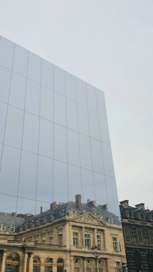 Reflection of a historic building with ornate architecture on the mirrored surface of a modern glass structure. The reflected building features classical elements such as columns and decorative moldings.