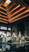 An elegant interior space featuring a high ceiling with layered geometric designs illuminated by warm orange and yellow lights. There are wooden furnishings, including chairs and tables. Decorative elements such as intricate wall carvings and large arched mirrors are present. Potted plants and ambient lighting from lamps on side tables enhance the decor.