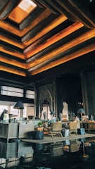 An elegant interior space featuring a high ceiling with layered geometric designs illuminated by warm orange and yellow lights. There are wooden furnishings, including chairs and tables. Decorative elements such as intricate wall carvings and large arched mirrors are present. Potted plants and ambient lighting from lamps on side tables enhance the decor.