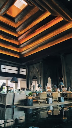 An elegant interior space featuring a high ceiling with layered geometric designs illuminated by warm orange and yellow lights. There are wooden furnishings, including chairs and tables. Decorative elements such as intricate wall carvings and large arched mirrors are present. Potted plants and ambient lighting from lamps on side tables enhance the decor.