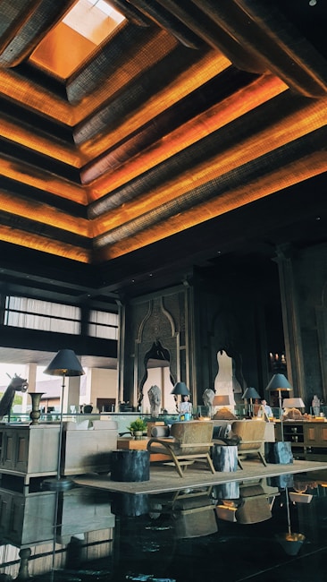 An elegant interior space featuring a high ceiling with layered geometric designs illuminated by warm orange and yellow lights. There are wooden furnishings, including chairs and tables. Decorative elements such as intricate wall carvings and large arched mirrors are present. Potted plants and ambient lighting from lamps on side tables enhance the decor.
