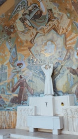 A detailed mosaic adorns a wall, depicting religious iconography and symbolism. In the center, a white stone statue of a robed figure stands with hands raised. The background is filled with abstract and geometric patterns in gold, blue, and earthy tones, featuring celestial and architectural elements. Below, a relief shows smaller figures gathered around an altar-like structure.