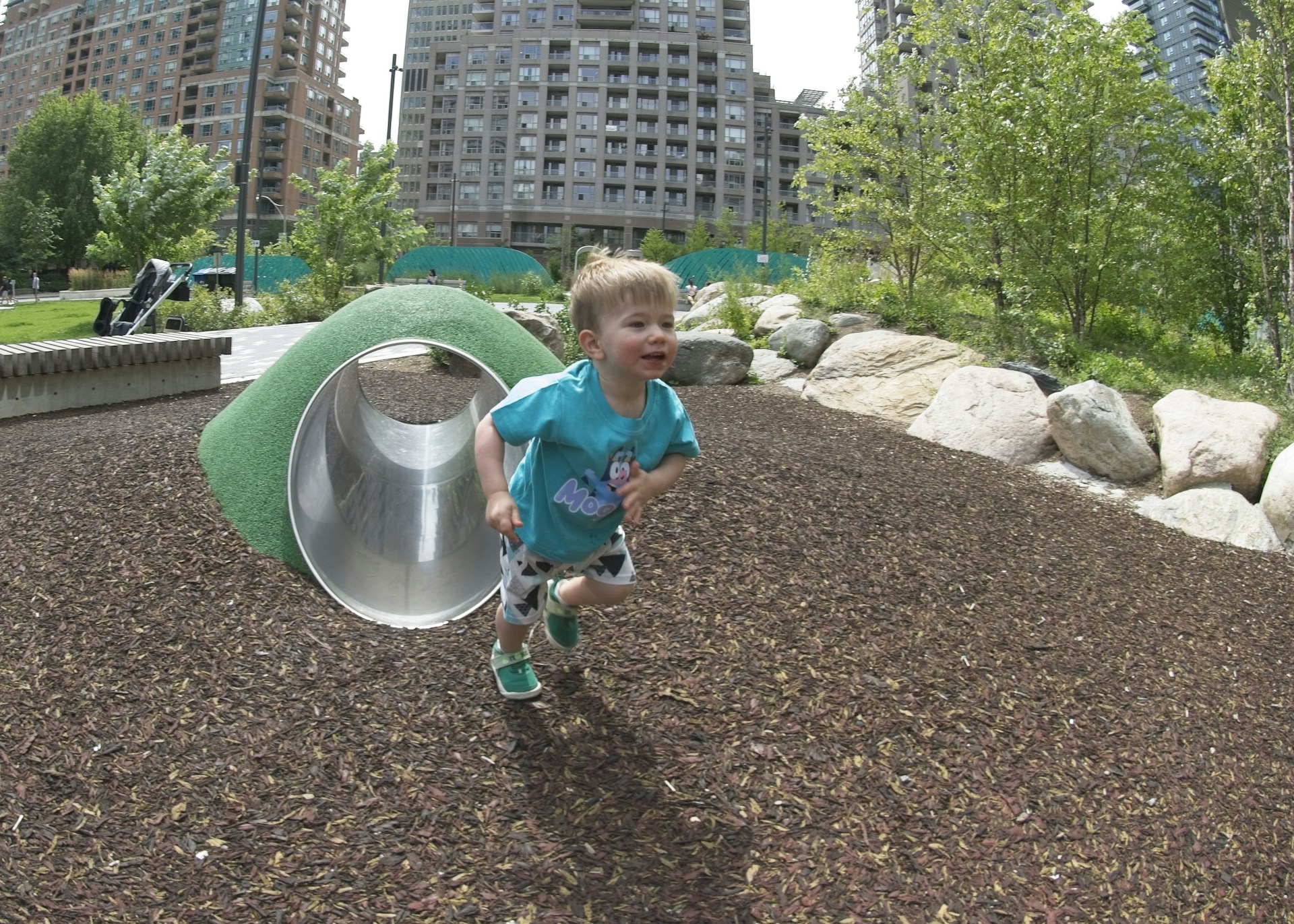 A toddler joyfully runs out of a small play tunnel in a park, surrounded by greenery and large buildings in the background. The child is wearing a blue shirt with cartoon graphics and patterned shorts. The playground is covered with mulch, and there are large rocks and bushes nearby.