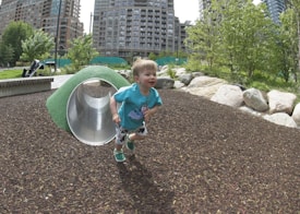A toddler joyfully runs out of a small play tunnel in a park, surrounded by greenery and large buildings in the background. The child is wearing a blue shirt with cartoon graphics and patterned shorts. The playground is covered with mulch, and there are large rocks and bushes nearby.