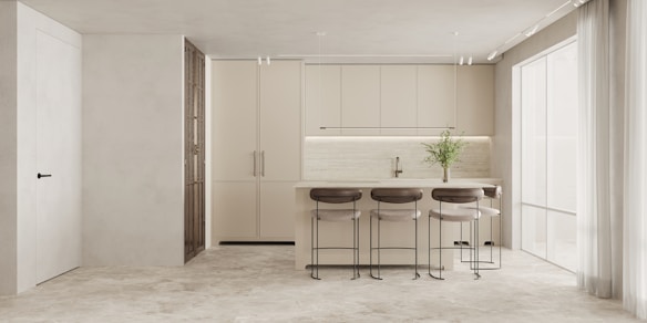 A modern kitchen with minimalist design, featuring a large island with four bar stools, sleek cabinetry in neutral tones, and a built-in sink. A vase with green plants adds a touch of nature, while large windows bring in natural light, enhancing the airy and open atmosphere.