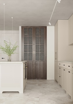 A modern kitchen with minimalist design features a central kitchen island and beige cabinets. The floor is made of light-colored tiles, and a vase with green foliage sits on the island. Natural light illuminates the room, enhancing the soft color palette. A set of large, dark wood paneled doors with intricate glass panes adds a focal point.