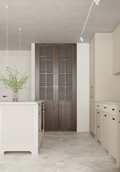 A modern kitchen with minimalist design features a central kitchen island and beige cabinets. The floor is made of light-colored tiles, and a vase with green foliage sits on the island. Natural light illuminates the room, enhancing the soft color palette. A set of large, dark wood paneled doors with intricate glass panes adds a focal point.