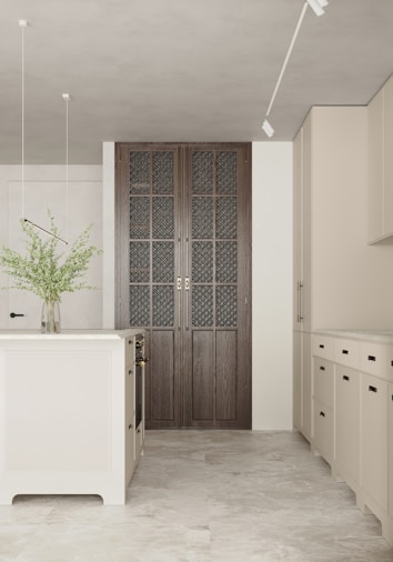 A modern kitchen with minimalist design features a central kitchen island and beige cabinets. The floor is made of light-colored tiles, and a vase with green foliage sits on the island. Natural light illuminates the room, enhancing the soft color palette. A set of large, dark wood paneled doors with intricate glass panes adds a focal point.