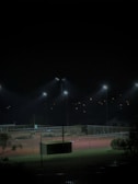 A dimly lit tennis court at night with several floodlights illuminating the area. The surrounding environment is mostly dark, with some trees and foliage visible in the foreground. Small lights are visible in the background, suggesting distant buildings or homes.