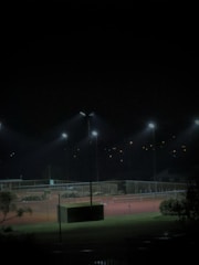A dimly lit tennis court at night with several floodlights illuminating the area. The surrounding environment is mostly dark, with some trees and foliage visible in the foreground. Small lights are visible in the background, suggesting distant buildings or homes.