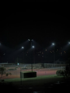 A dimly lit tennis court at night with several floodlights illuminating the area. The surrounding environment is mostly dark, with some trees and foliage visible in the foreground. Small lights are visible in the background, suggesting distant buildings or homes.