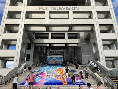 People are walking on a staircase with colorful artwork depicting vibrant imagery, possibly an advertisement or promotional art. The artwork is situated at the base of a large modern building, likely a television station due to the signage 'Fuji Television' at the top. The structure is composed of a grid-like pattern with large rectangular sections.