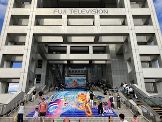People are walking on a staircase with colorful artwork depicting vibrant imagery, possibly an advertisement or promotional art. The artwork is situated at the base of a large modern building, likely a television station due to the signage 'Fuji Television' at the top. The structure is composed of a grid-like pattern with large rectangular sections.