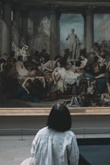 A person is observing a large, classical painting in an art gallery. The artwork features numerous figures in a dramatic scene, possibly inspired by mythology or history. The painting includes various human forms, some draped in robes, and a prominent statue in the background.