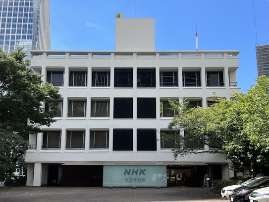 A white multi-story building with a flat roof and rectangular windows. The facade is symmetrical, featuring black sunshades on some windows. The entrance has a sign with the letters 'NHK' and is surrounded by trees. Cars are parked in front, and the sky is clear and blue.