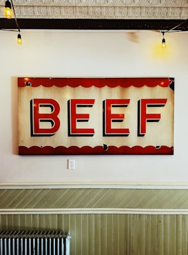 A vintage-style sign with bold red letters spelling 'BEEF' against a cream background, bordered by scalloped red edging. The sign is displayed on a white wall, and there are exposed hanging bulbs from the ceiling, contributing to a rustic atmosphere. Below the sign, a green wainscoting and a radiator are visible.