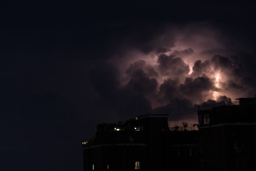 Dark storm clouds illuminated by flashes of lightning, hovering above the silhouette of urban buildings.