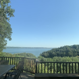 A serene landscape with a clear blue sky over a large body of water, surrounded by lush green trees. A wooden deck with outdoor furniture overlooks the scene, providing a peaceful spot for relaxation.