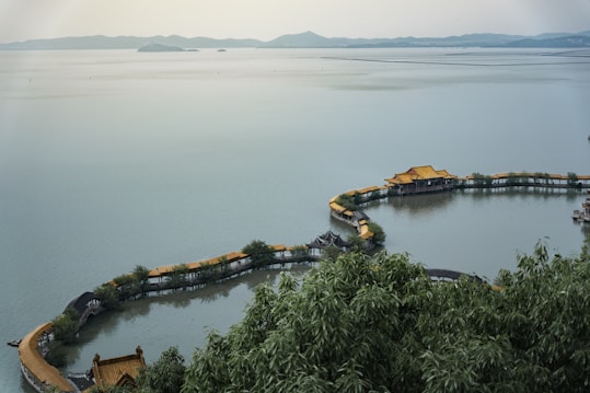 A serene landscape featuring a large body of calm water with a circular pathway lined with traditional Chinese-style rooftops and greenery extending into the water. The distant background includes hazy mountains under an overcast sky, creating a tranquil and peaceful setting.