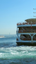 A ferryboat with lifebuoys along its railing is seen in the foreground, its hull labeled with 'SH-Beykoz Istanbul.' The water shows vibrant greenish-blue swirls indicating movement. In the background, a body of water extends towards a suspension bridge under a clear blue sky. Another smaller boat is present in the distance.