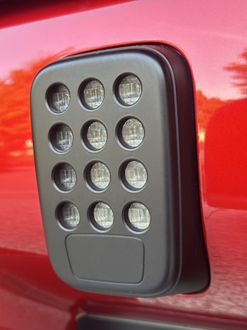 A close-up view of a rectangular black plastic component with multiple circular clear lenses arranged in a grid pattern. The component is mounted on a glossy red surface, likely part of a vehicle.
