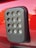 A close-up view of a rectangular black plastic component with multiple circular clear lenses arranged in a grid pattern. The component is mounted on a glossy red surface, likely part of a vehicle.