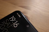 A close-up view of the top corner of a smartphone, displaying the time 19:16, the day SAT, and the date 20/10. The phone screen shows two network providers, Jio and BSNL, in the top right corner. The phone is placed on a wooden surface, with a smooth and slightly reflective finish.