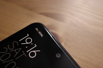 A close-up view of the top corner of a smartphone, displaying the time 19:16, the day SAT, and the date 20/10. The phone screen shows two network providers, Jio and BSNL, in the top right corner. The phone is placed on a wooden surface, with a smooth and slightly reflective finish.