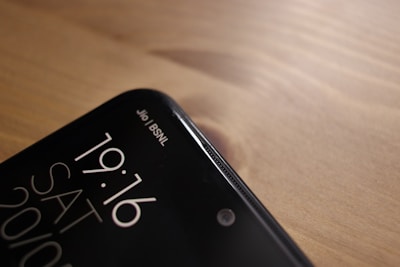 A close-up view of the top corner of a smartphone, displaying the time 19:16, the day SAT, and the date 20/10. The phone screen shows two network providers, Jio and BSNL, in the top right corner. The phone is placed on a wooden surface, with a smooth and slightly reflective finish.