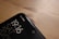 A close-up view of the top corner of a smartphone, displaying the time 19:16, the day SAT, and the date 20/10. The phone screen shows two network providers, Jio and BSNL, in the top right corner. The phone is placed on a wooden surface, with a smooth and slightly reflective finish.